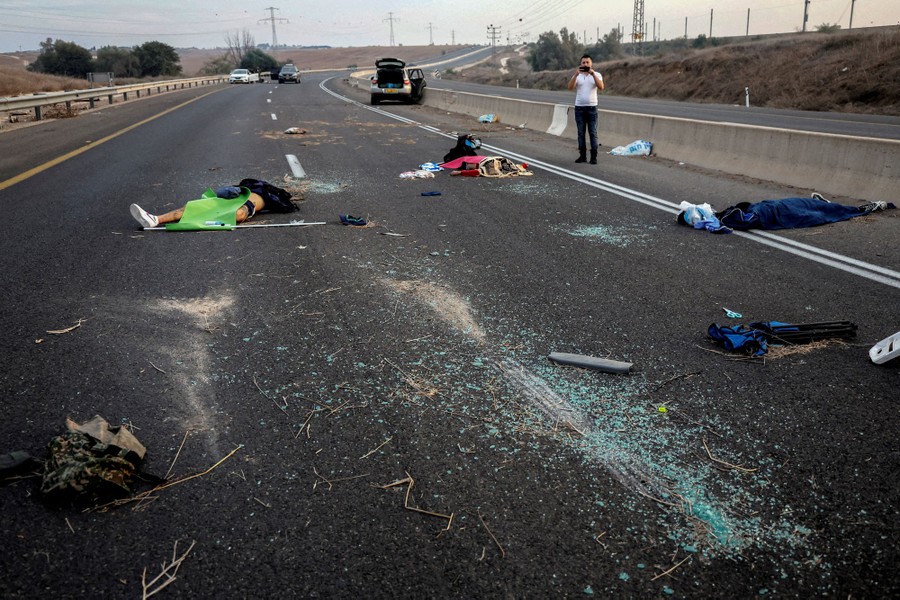 A man takes a photo of several dead people strewn across a road alongside broken glass and debris.