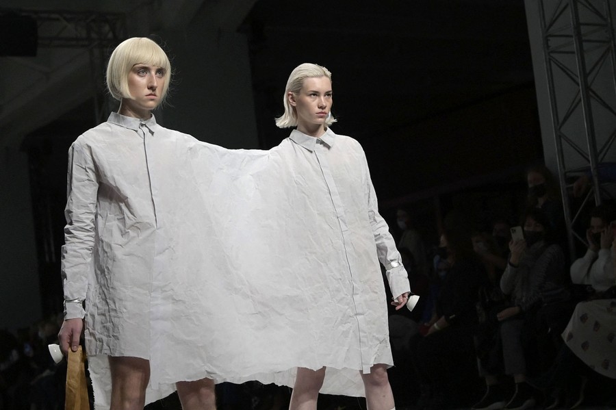 A pair of models wear a single, wide, connected shirt dress on a runway.