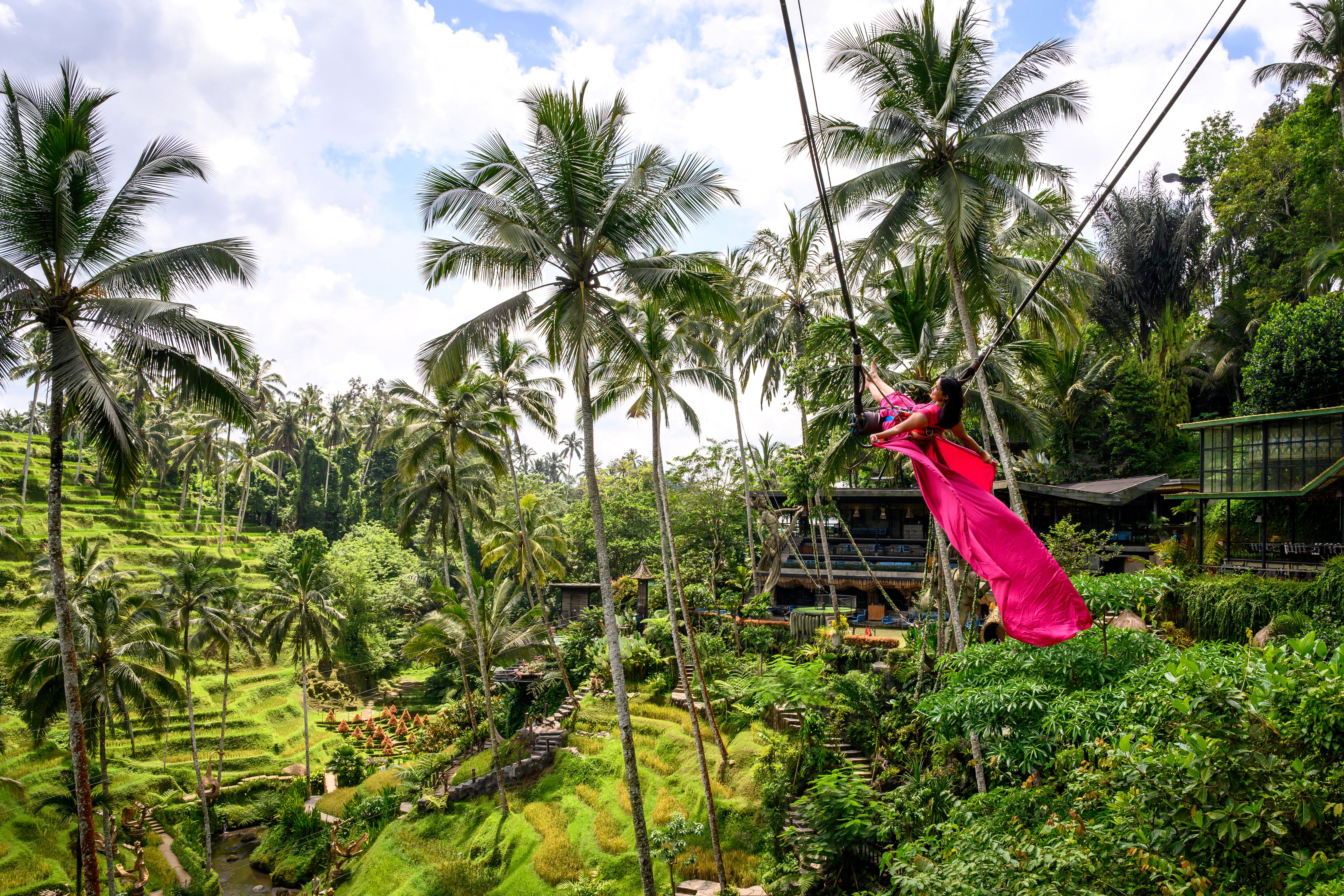 A woman in a long red dress poses for photographs on a giant swing above a lush valley.
