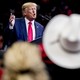 President Trump speaks into a microphone at a campaign rally as supporters, one wearing a white cowboy hat, look on.