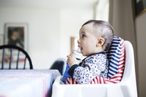 A baby sits in a high chair holding a bottle.