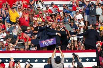 A photograph of Donald Trump surrounded by Secret Service agents on a stage; people in the crowd looking on raise their arms and hold up their cameras.