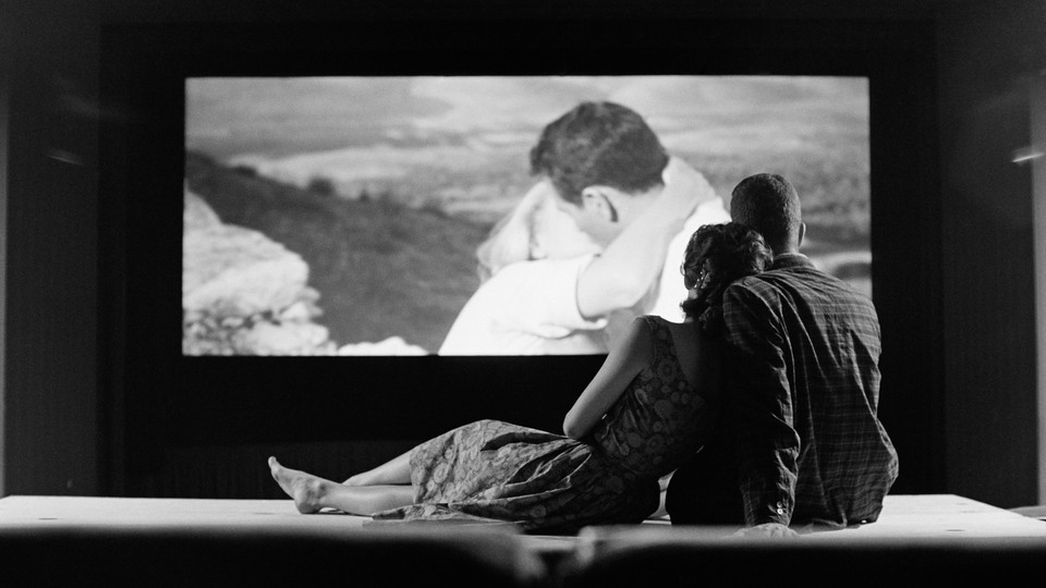A black-and-white photo of a heterosexual couple watching another heterosexual couple kiss on a big screen
