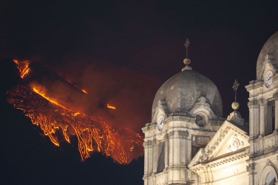 Lava is seen flowing down a slope behind an illuminated church at night.