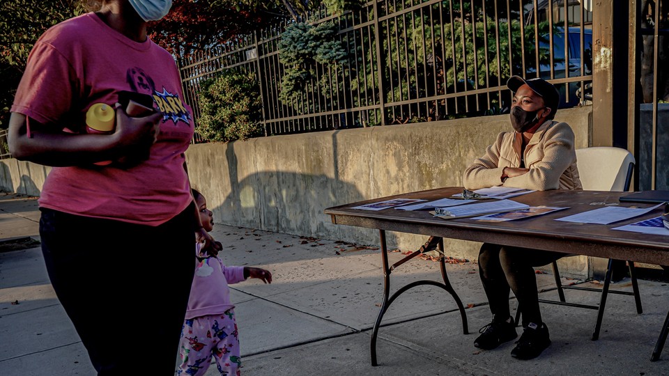 A woman and child walk past a voter-registration table in Brooklyn. Another woman is sitting at the table. Both adults are wearing masks.