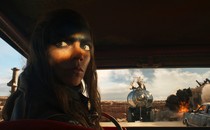 A film still showing Anya Taylor-Joy as the character Furiosa looking backward while sitting in a car; other cars, and an explosion, can be seen through the windshield in the desert landscape behind her.