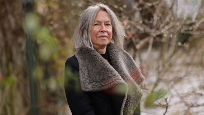 A portrait photo of Louise Glück in black coat standing outside among bare branches