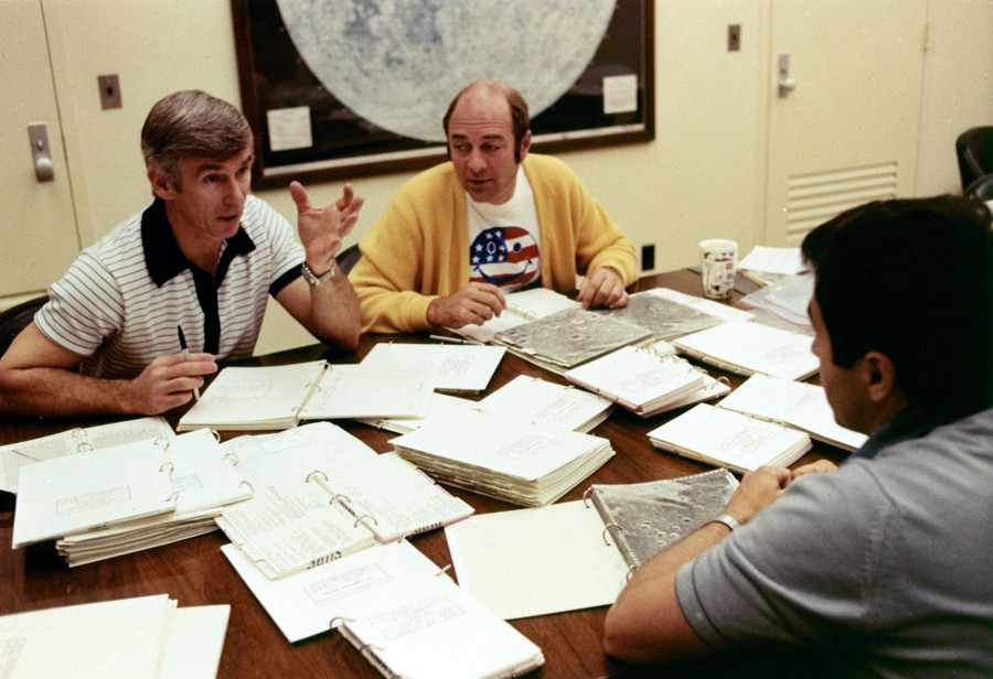 Three men sit at a table covered in notebooks and maps, having a discussion.