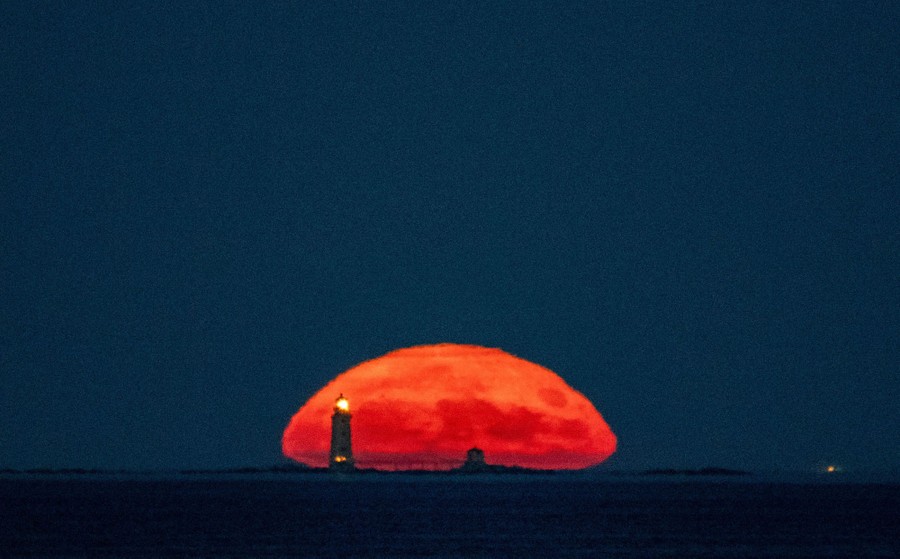 A reddish moon is seen near the horizon, slightly squashed by atmospheric distortion, behind a lighthouse on a small island.