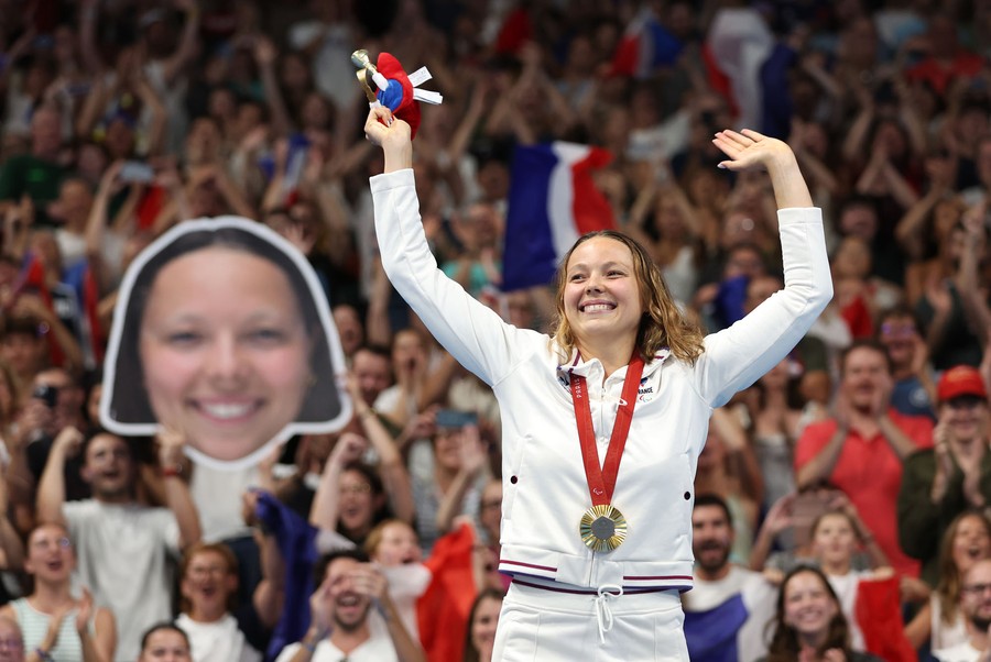 An athlete waves to the crowd at a medal ceremony, as fans hold up a large cutout of her face in the background.