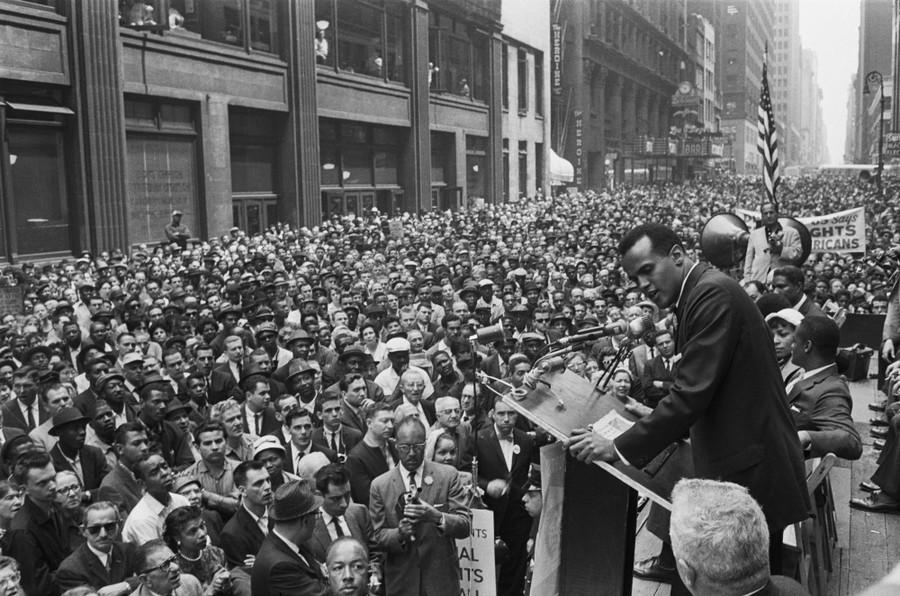 Harry Belafonte stands at a lectern, singing in front of a large crowd of people standing in a street.
