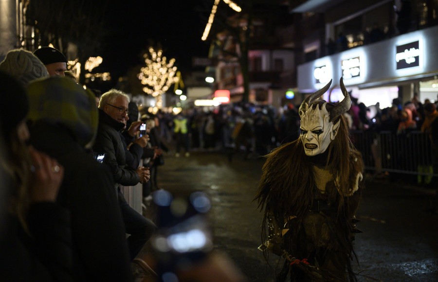 A crowd gathers in a village street, watching as a performer in a demon mask passes by.