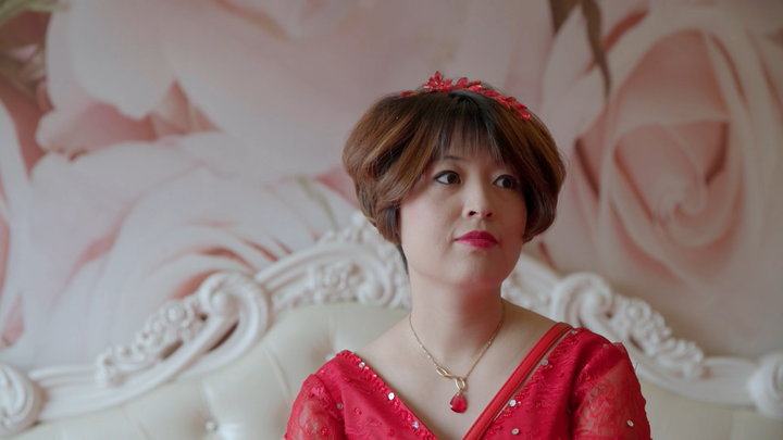 Closeup of Gai Qi, one of the women in the documentary, wearing a red dress against a pink backdrop.