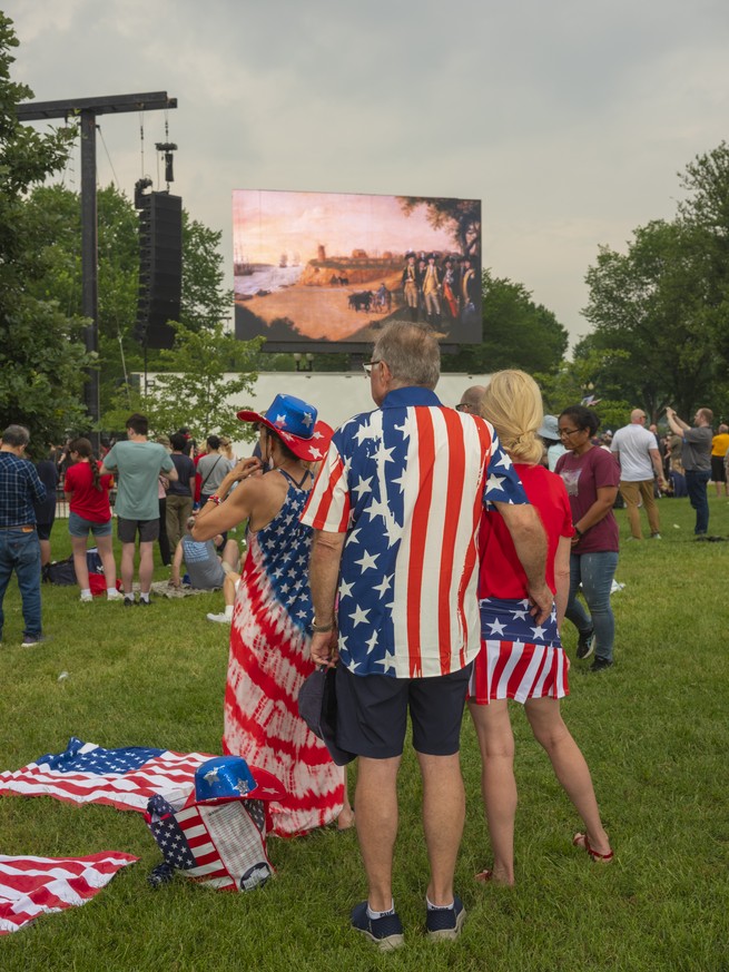 Picture of parade-goers in patriotic ensembles standing near a screen showing a video about the Army’s history. 
