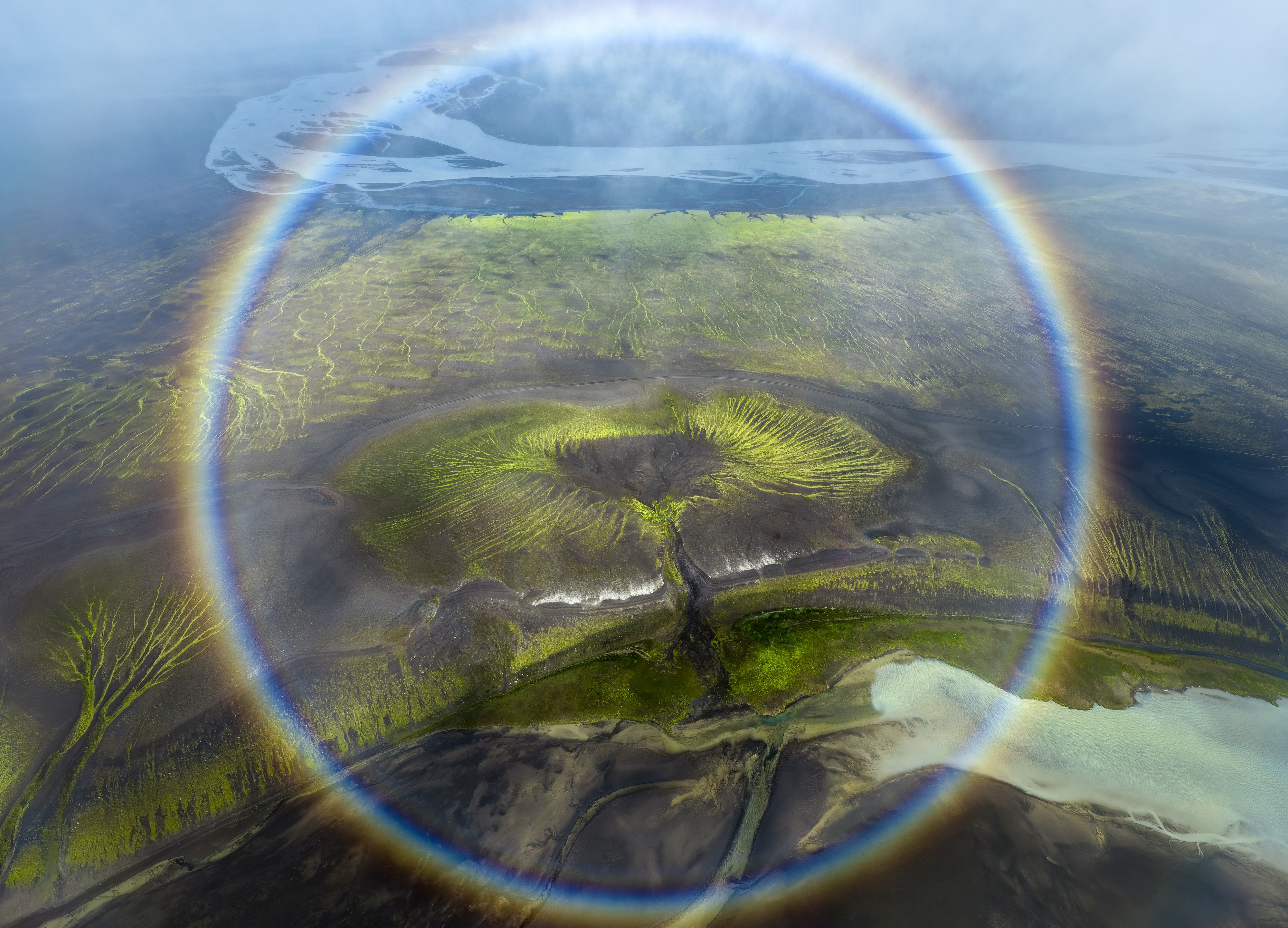 An aerial view of a green river plain, seen through a circular rainbow