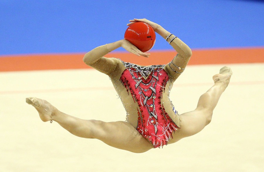 A gymnast leaps while holding a ball, her head thrown so far back it appears to be missing, seen from this angle.