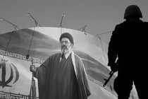Black-and-white photograph of an armed soldier standing in front of a mural of Mojtaba Khamenei holding a rifle next to the Iranian flag