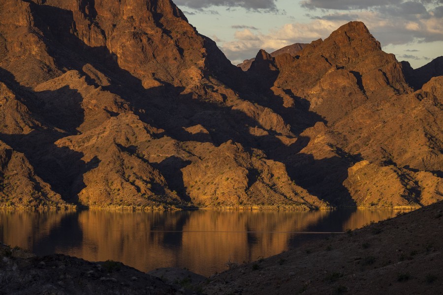 Low sunlight shines on rugged mountains above a reservoir.