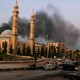 Photo of street view in Aleppo with smoke billowing above buildings and wrecked bombed cars in the foreground.