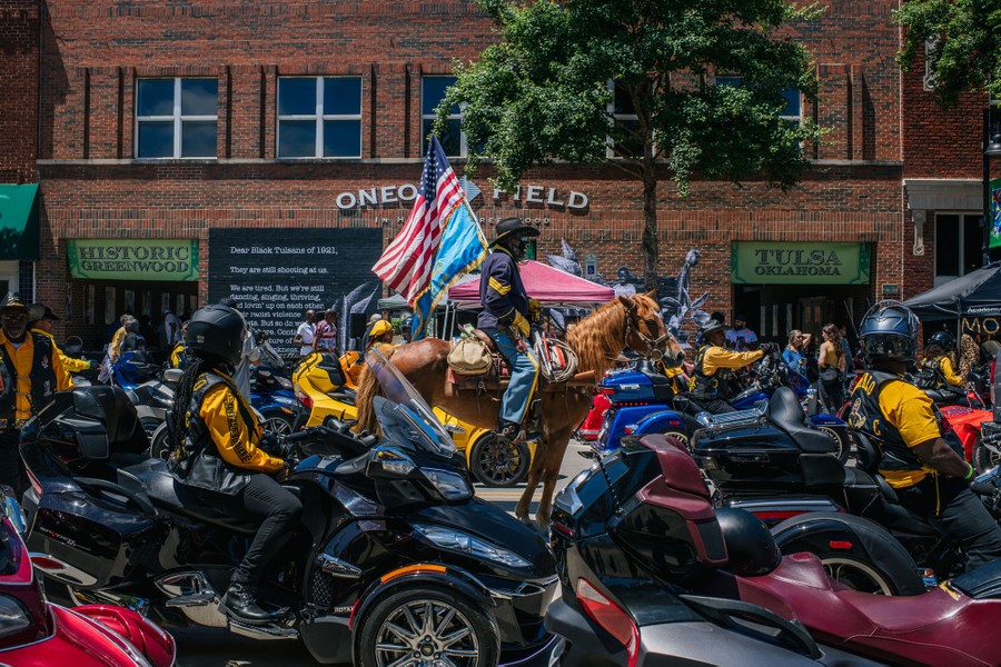 Several people on motorcycles and one on horseback (carrying an American flag) ride together in a street.