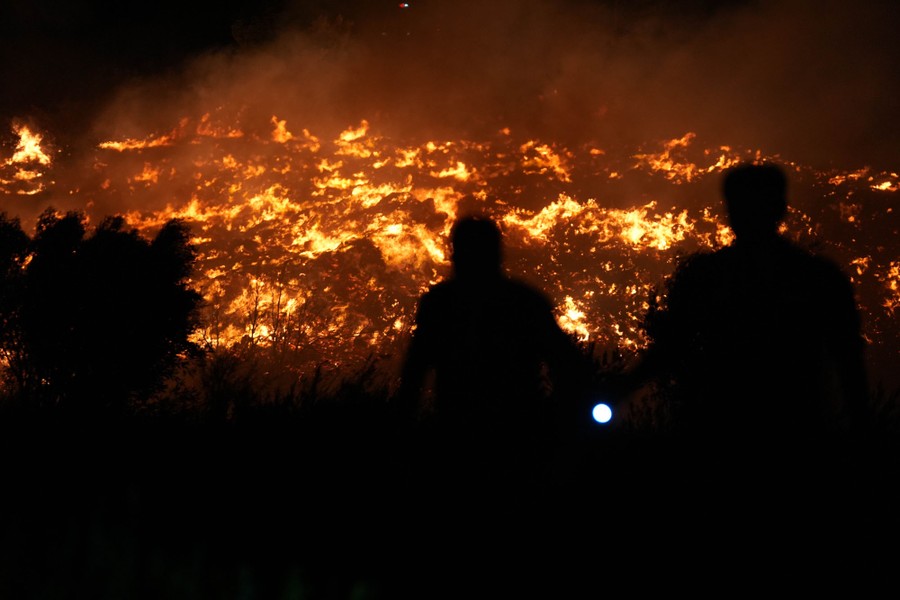 Two people are silhouetted in front of fires burning on a hillside.