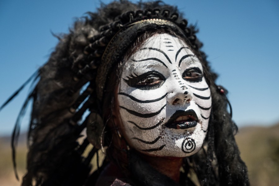 A woman poses for a photograph wearing a headdress; her face is painted in white makeup and decorated with black stripes.