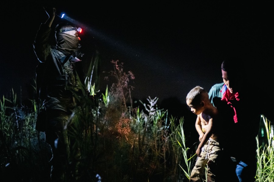 A member of the U.S. National Guard points a flashlight at a person carrying a child through reeds on a river bank at night.