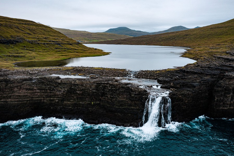An aerial view of a steep waterfall emptying a cliffside lake into the ocean.
