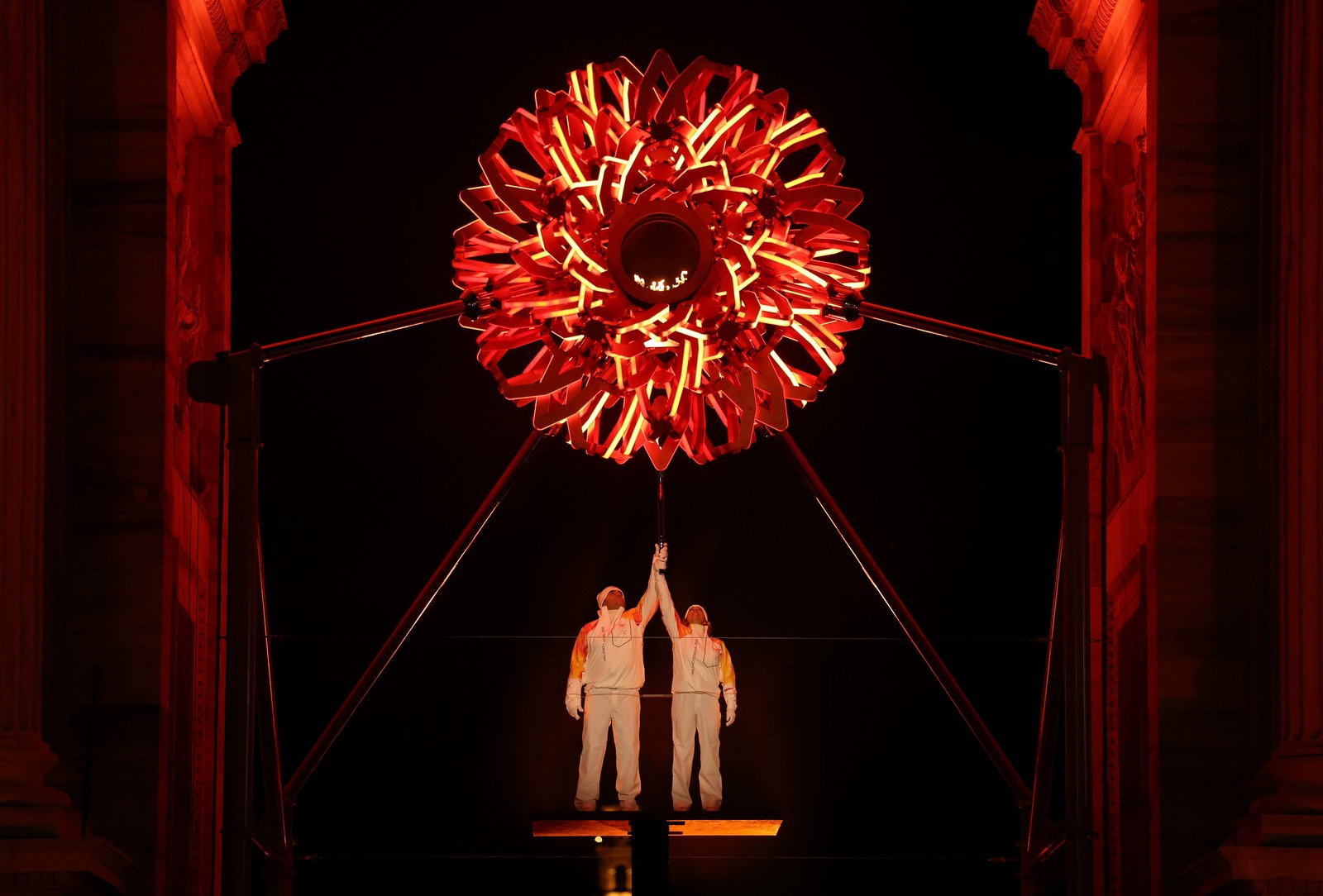 Two athletes jointly hold a torch aloft, standing beneath a tall arch, lighting the ornate spherical Olympic cauldron above them.