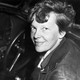 Black and white photograph of Amelia Earhart in the cockpit of a plane, looking straight into the camera with a smile