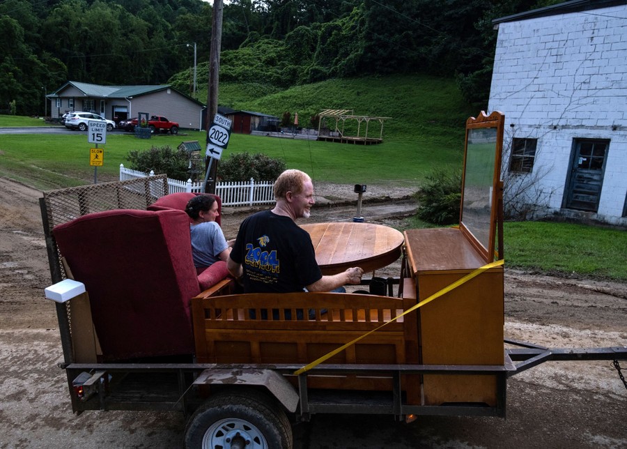 Two people sit among furniture being moved on a trailer on a mud-covered road.