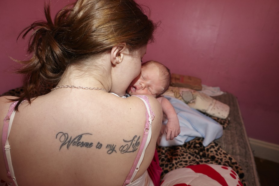 A woman seen from the back with a tattoo reading "Welcome to my Life" in a pink room holding a baby.