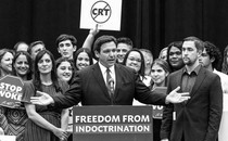A black-and-white photograph of Ron DeSantis at a podium with his arms outstretched and a group of people standing around him holding signs against "critical race theory."