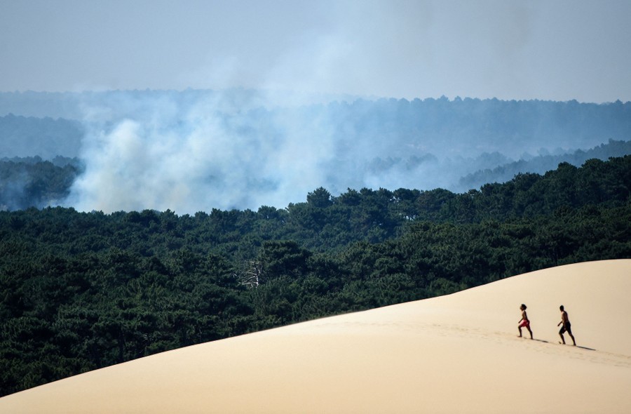 Two people walk on a large sand dune, with smoke from a forest fire billowing below them.