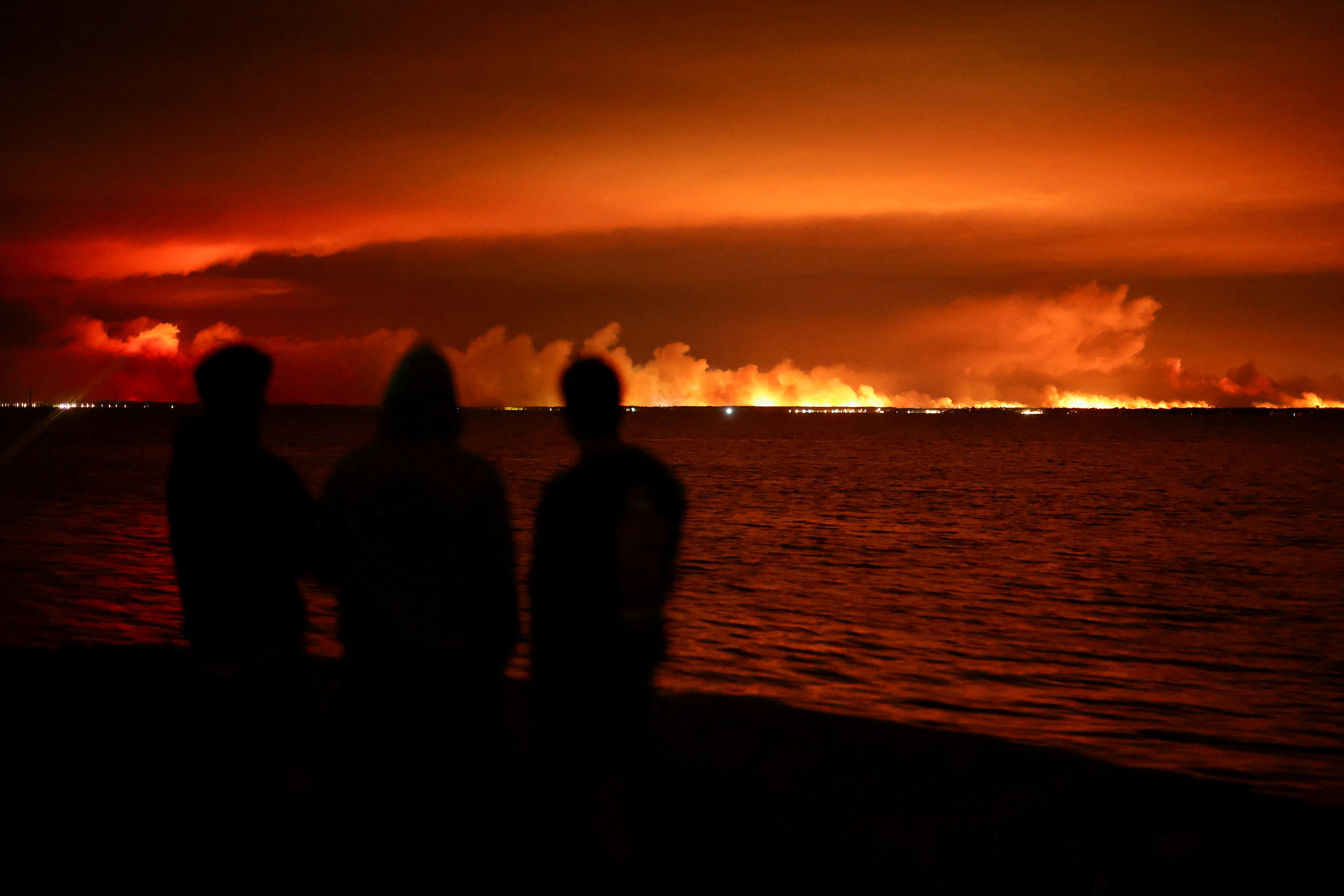 Several people watch a wildfire burn, seen across a body of water, at night.