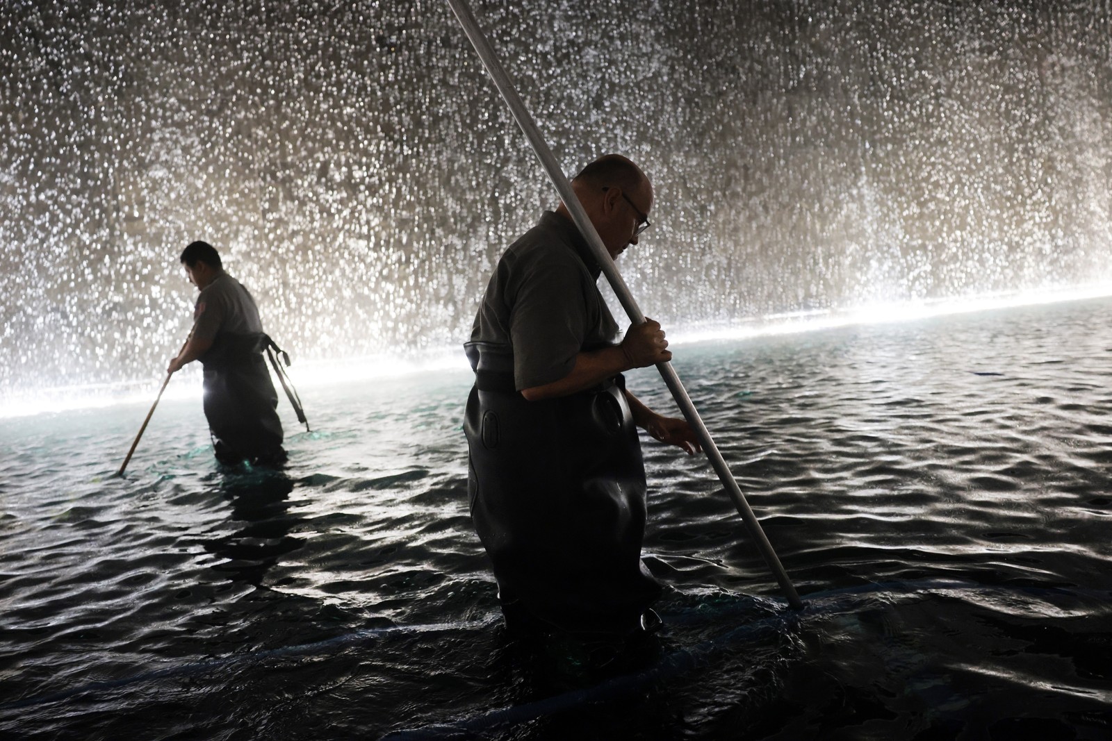 Two people wearing waders clean a memorial pool at night, with a waterfall behind them.