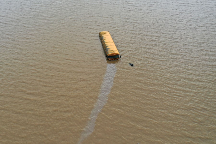 A small oil slick trails away from a submerged school bus in a flooded area.