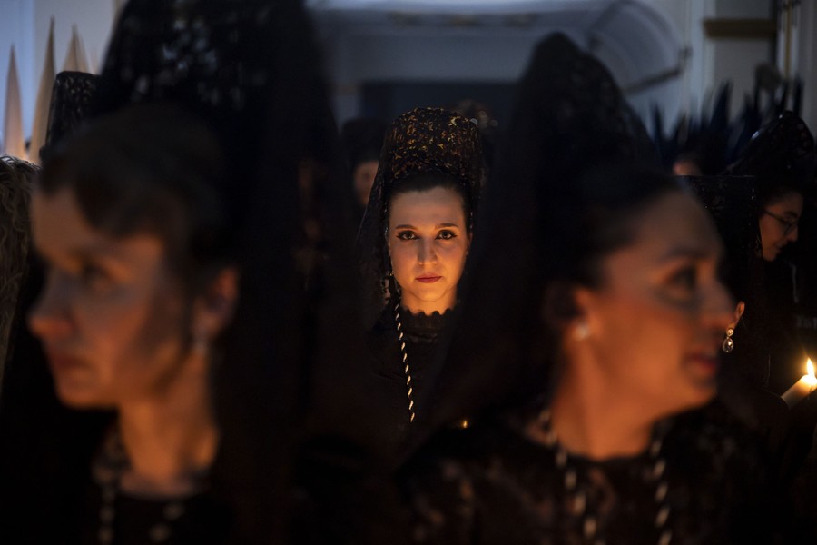 A group of women wearing traditional clothing, holding candles, gather in a room, preparing for a procession.