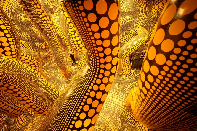A person walks through an exhibit of a yellow sculpture with black dots