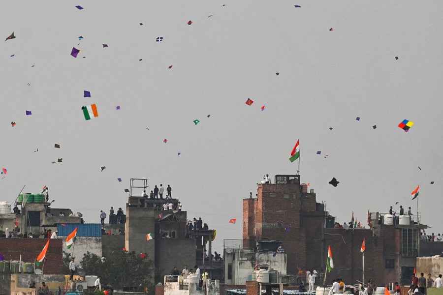 Dozens of kites appear in the sky above buildings, where people stand on rooftops.