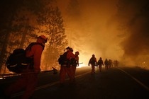 A line of firefighters walks down a road as flames loom in the background.