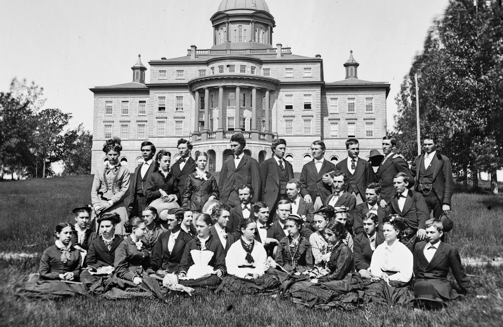 Thirty-five young people in formal clothing pose outside of a tall school building