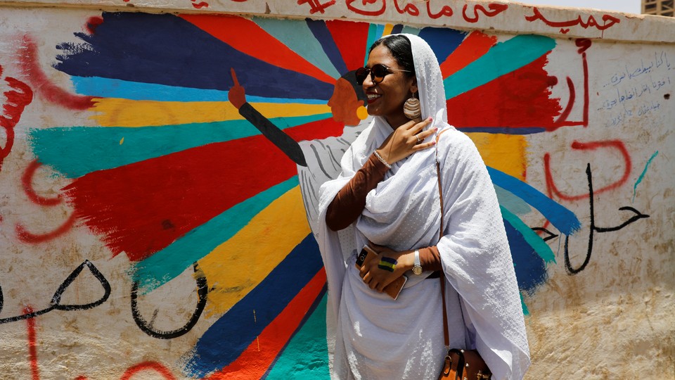 Alaa Salah, dressed all in white, stands in front of a mural of herself.