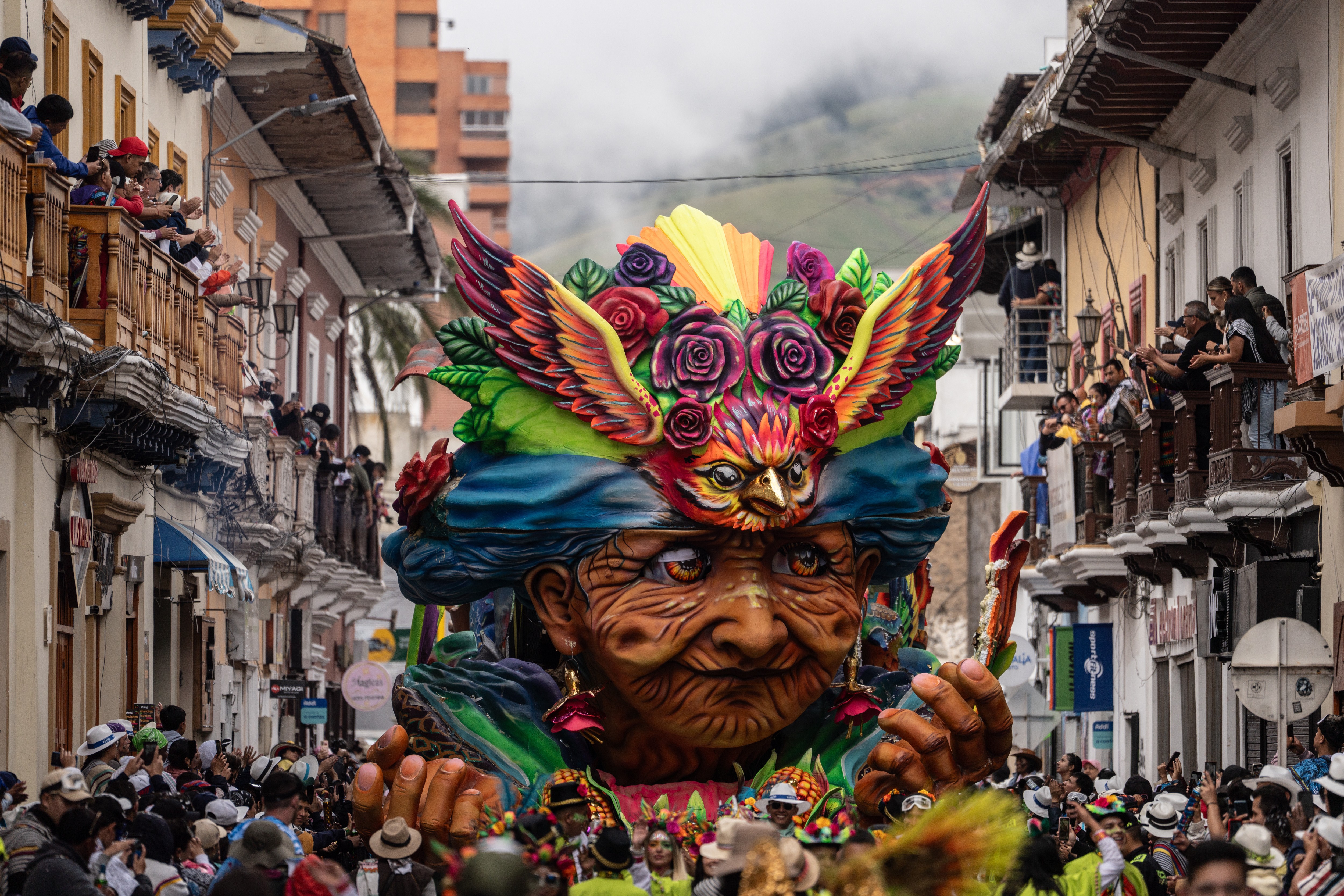 A crowd watches from the sides of a street and balconies as a parade float shaped like a giant head with a colorful headdress passes by.