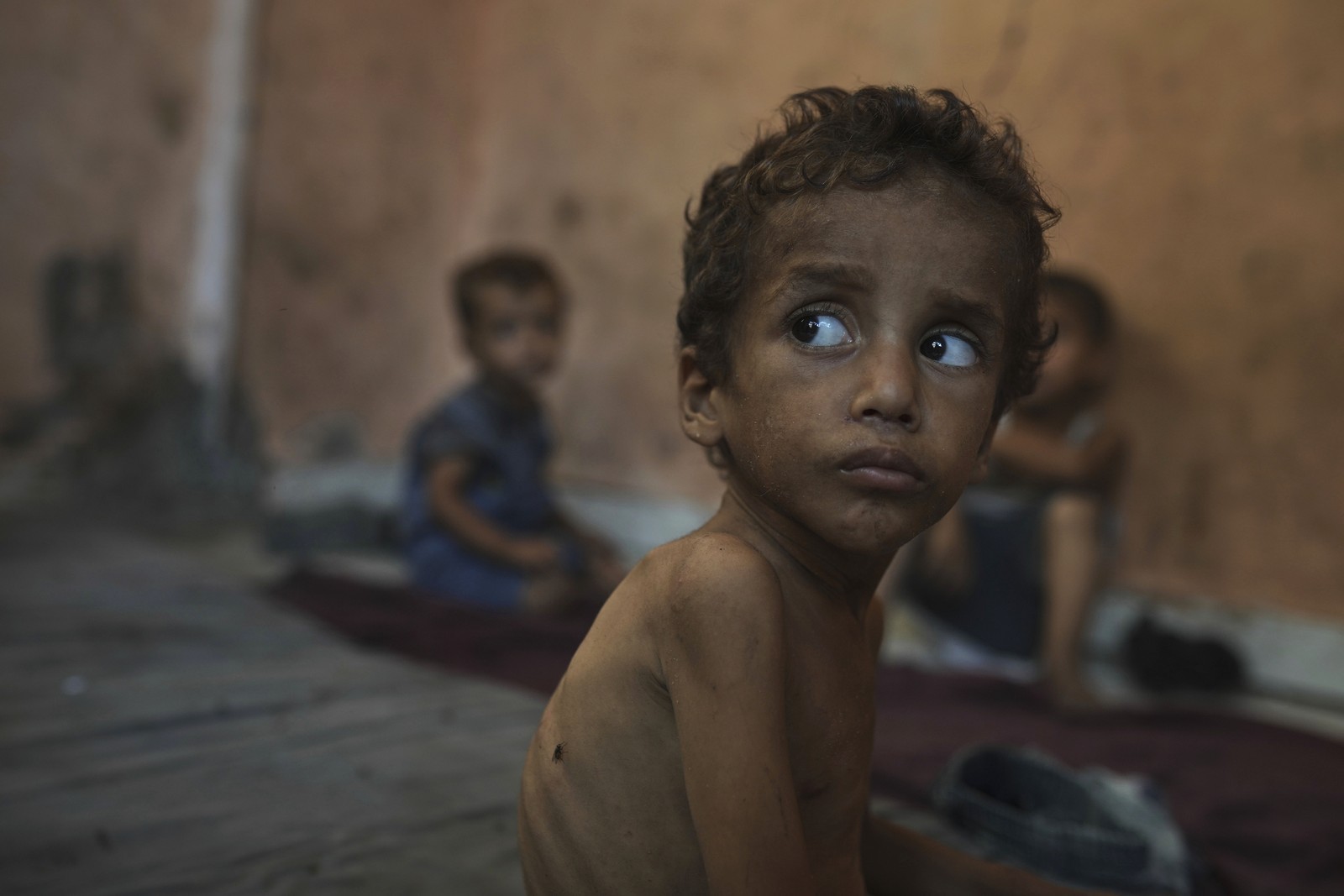 A close view of a young boy who is very undernourished, sitting on the floor near to other children.