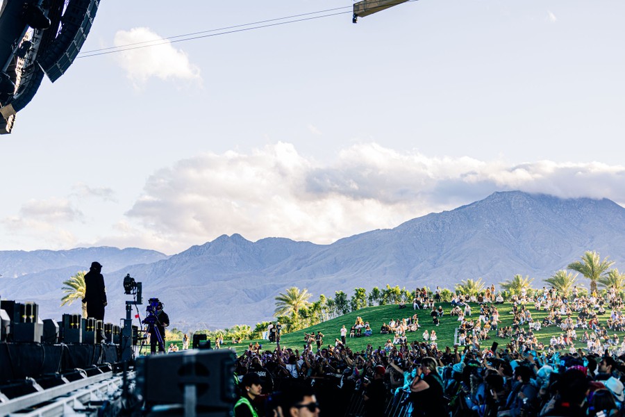 Concertgoers stand and sit on a shallow grassy hill while watching a performance.