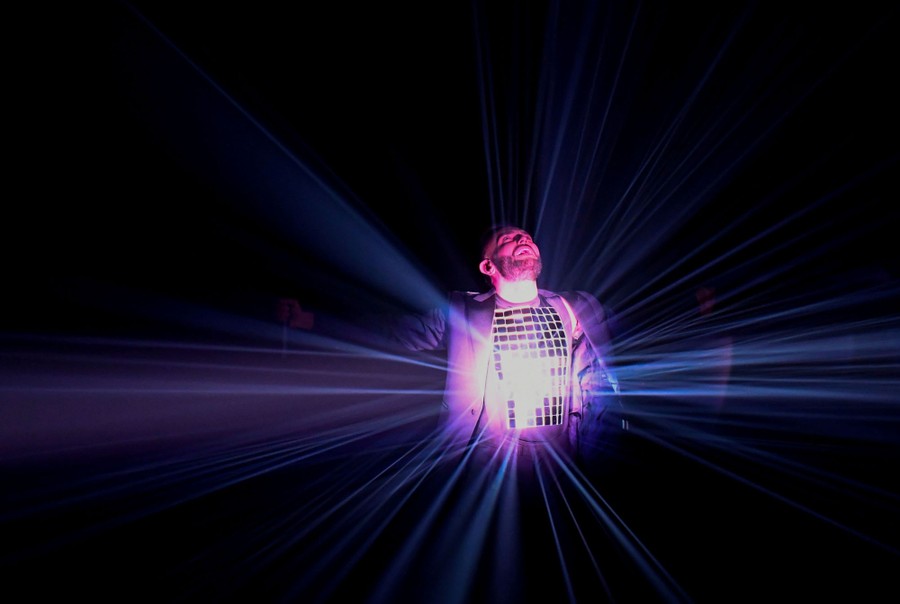 A performer stands on a stage wearing a mirrored vest that is lit by a spotlight.