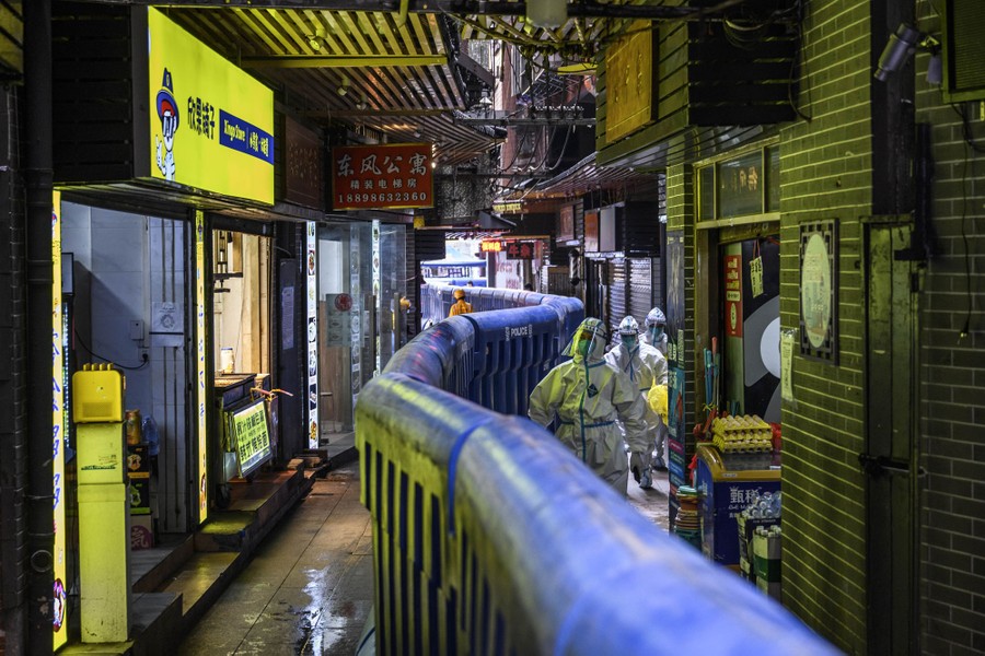 Several people in protective clothing and masks walk beside a temporary barrier wall that runs down the sidewalk of a city marketplace.