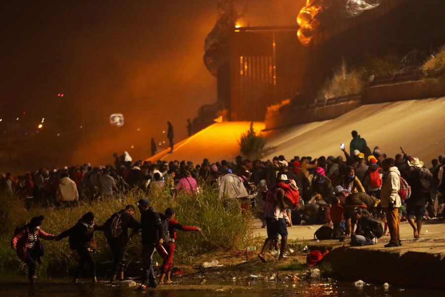 A large crowd of people gather on a riverbank, alongside a tall border fence.