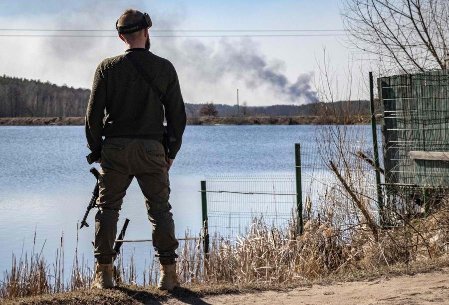 A man holding a rifle looks across a body of water toward a line of black smoke rising in the distance.